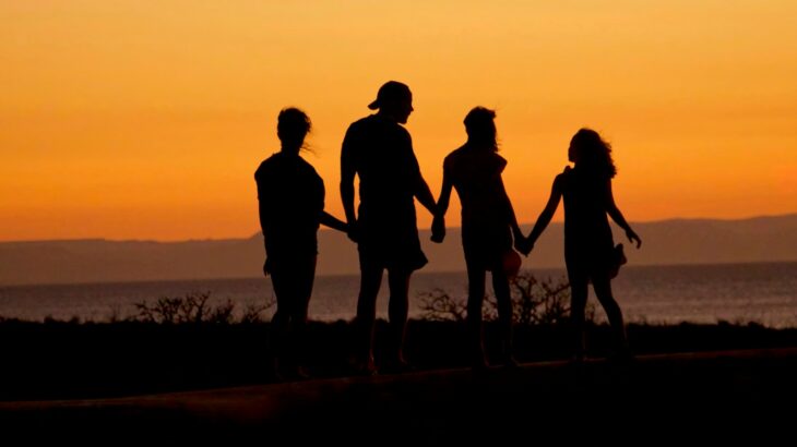 A family in silhouette holding hands against a warm orange sky by the sea