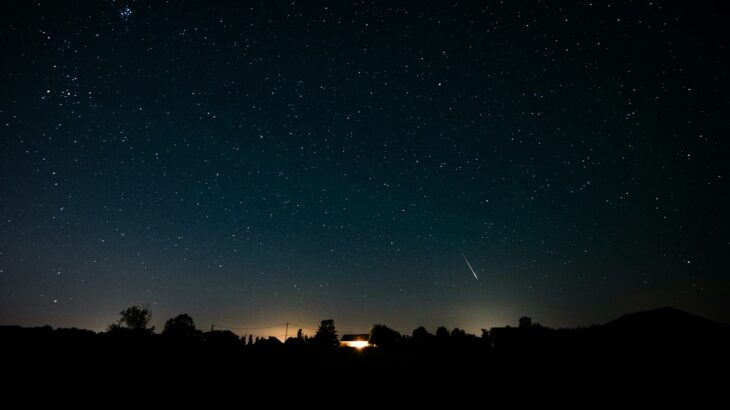 A starry night sky with a shooting star over a silhouetted treeline and house